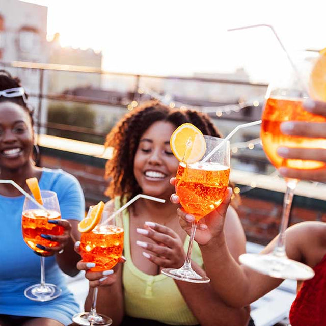 An outdoor photo of a group of women, each one of them raising a wine glass of orange fizzy drink.