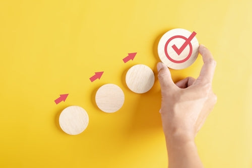 Hand places a wooden disc with a red check mark at the end of a row of discs linked by rising red arrows, on yellow.