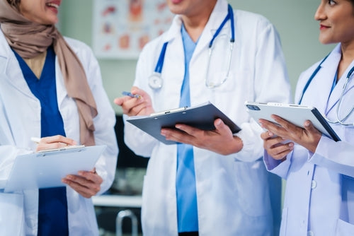 Three doctors in white coats with stethoscopes confer while holding clipboards and a tablet.