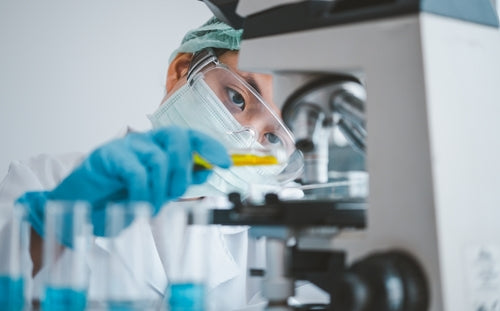 Masked scientist in safety goggles and blue gloves examines a vial of yellow liquid beside a microscope.